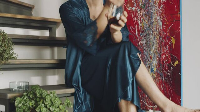 Handheld camera shot of barefoot transgender man in silk robe sitting on wooden stairs at home, drinking alcohol cocktail and smoking cigarette