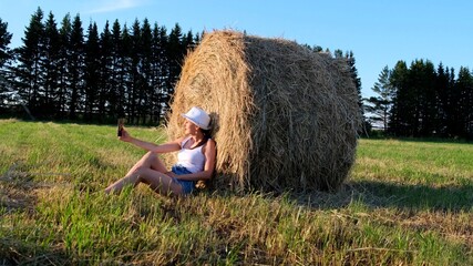 Naklejka na ściany i meble young female makes a selfie on a smartphone near a haystack and talks on video communication. Rural area. freedom concept