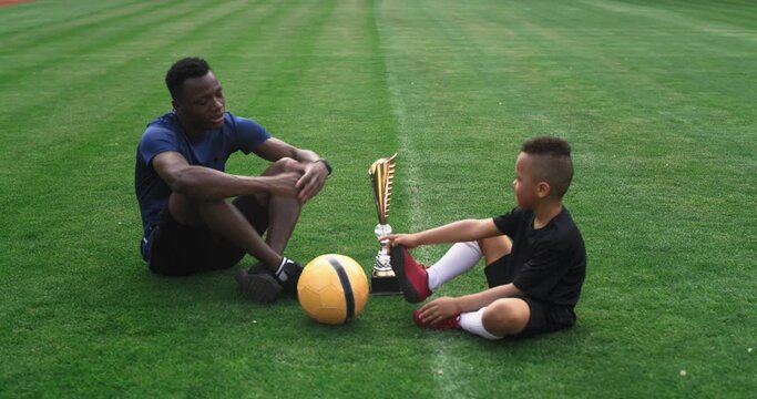 Diverse father and son talking near golden cup on football pitch