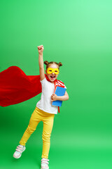 happy smiling schoolgirl in a red cape and a superhero mask and with a school book in her hands,runs and jumps on the green background of the studio. The child is ready for successful learning. © skif