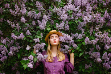 Portrait of a young beautiful woman in a straw hat near a blooming lilac bush. The girl looks away dreamily. Copy, space for the text.