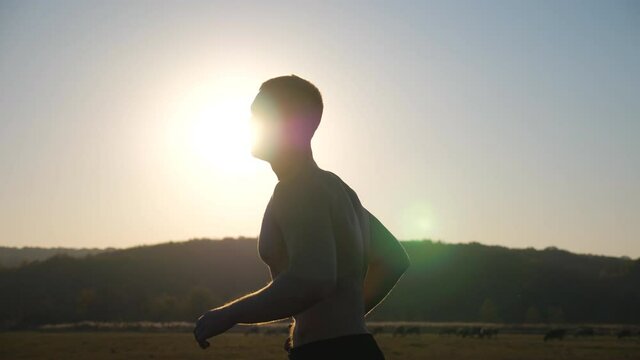 Young muscular man running through field with sunset at background. Strong and hardy sportsman running outdoor. Athletic soldier training at nature. Concept of healthy active lifestyle. Side view