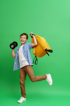happy emotional schoolboy in fashionable clothes with dumbbells and a backpack in his hands shows his strength on a green background in the studio. Cheerful smiling boy, dynamic image.