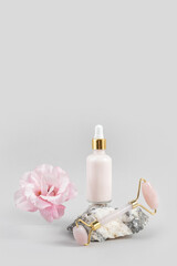 Crystal rose quartz facial roller, cosmetic dropper bottle on stone and beautiful flower on grey background. Facial massage for natural lifting, Beauty concept Front view