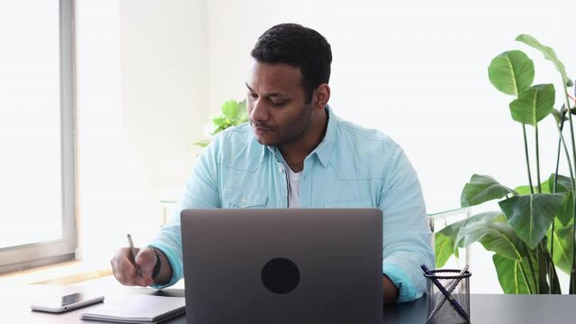Busy multi-ethnic Indian businessman working using laptop sitting at the desk in the office. Young man takes notes in a notebook for new startup