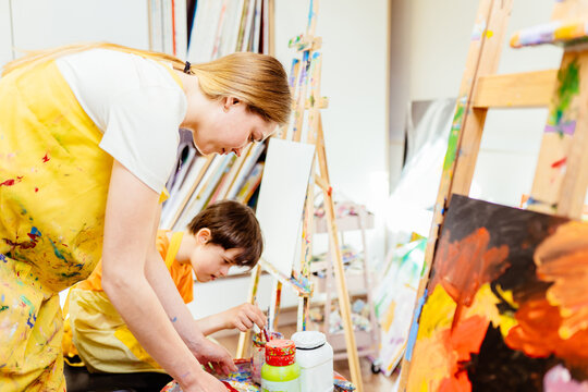 Education And Special Child Concept. Nursery Or Female Teacher Sitting With Boy Child Down Syndrome Drawing Drawing On Canvas On The Easel In The Art Class At School.