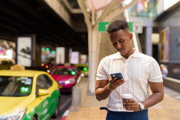 Portrait of young African businessman wearing casual clothes while using mobile phone and holding coffee cup at taxi station in city