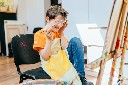 Education and special child concept. Cute positive happy boy with down syndrome laughing while sitting in a chair and drawing with paintbrush indoor.