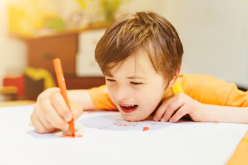 Happy boy child Down Syndrome in orange shirt drawing with pencil on a large white sheet of paper...