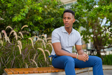 Portrait of young African businessman wearing casual clothes and sitting at park bench while thinking