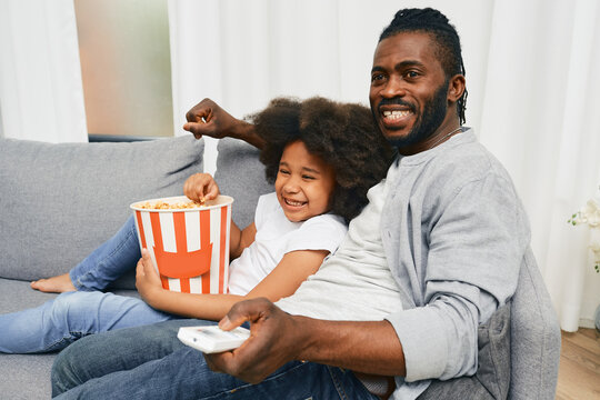 African American father with his little daughter hugging and watching cinema together and eating popcorn from a large striped bucket. Father's day