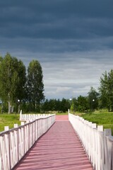 pink wooden footbridge with white railing on background of green forest and cloudy sky
