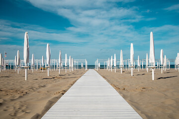 Riviera Romagnola spring view. Almost summer, clue skies, white walking path, sunbeds and umbrellas ready for season opening. Italian vintage riviera beach on the adriatic coast.  - obrazy, fototapety, plakaty
