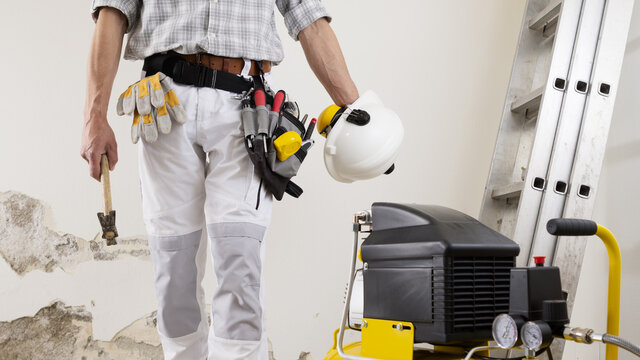 construction man worker wearing a tool bag belt, helmet with headphones and hammer in his hands, gloves in your pocket, compressor  and ladder on white wall background