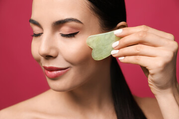 Beautiful young woman doing facial massage with gua sha tool on pink background, closeup
