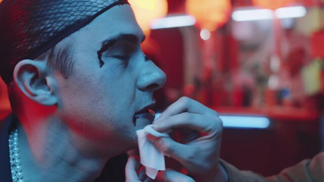 Close up shot of young woman cleaning blood from face of unhappy transgender male friend as he taking off wig and sitting in hairnet after bar brawl