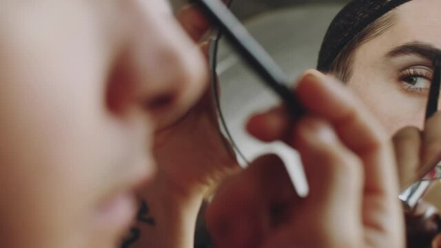 Over the shoulder shot of handsome transgender man in hairnet applying eyeshadow with brush and then looking at his mirror reflection while doing makeup in morning