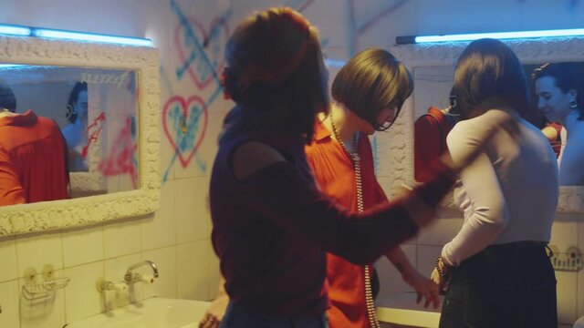 Joyous transgender man in female outfit smoking cigarette and laughing while chatting with two girlfriends in nightclub bathroom