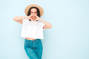 Portrait of young beautiful smiling female in trendy summer hipster  clothes. Sexy carefree woman posing near blue wall in studio. Positive model having fun in sunglasses and hat. Makes heart sign