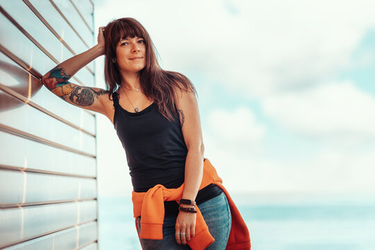 A young beautiful woman with tattoos poses, leaning against a wooden wall. In the background, the sky with clouds and the sea. Copy space