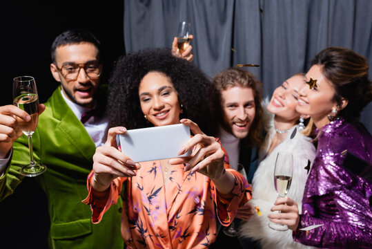 interracial friends in bright clothes taking selfie on smartphone near grey curtain on black background.