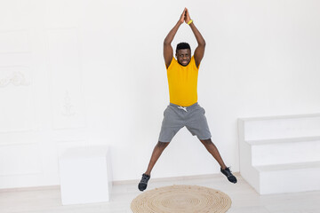Athletic Afro American male jumping, warming up during sport workout at home. Energetic sporty man in yellow sportswear doing jumping exercise, motivate to healthy lifestyle, training concept - Powered by Adobe