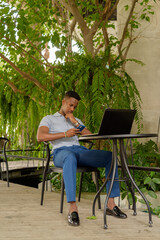 Portrait of young African businessman wearing casual clothes and sitting at coffee shop while using laptop computer and mobile phone while social distancing