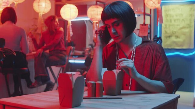 Transgender man in wig and female outfit talking on mobile phone while sitting at table in Chinese restaurant with noodles and drink