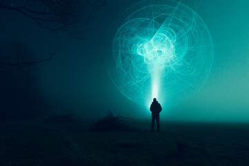 A science fiction concept. Of a man with a torch looking at an alien UFO in the sky, in a field on...