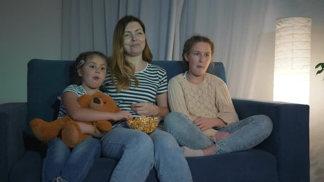 Happy family watching TV at home. Mom and kids are watching tv with popcorn. Happy family watching movie at home. Mom and children are eating popcorn on the couch. Happy family watching movie