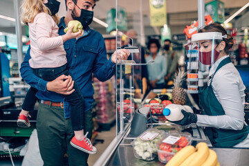 Beautiful smiling cashier working at a grocery store. Father is holding his daughter at market's checkout while paying by debit card. They are all wearing face protective masks.