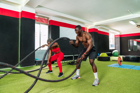 Black personal trainer coaching a bodybuilding woman to perform crossfit battle ropes exercise in the fitness gym. Workout training coach business concept