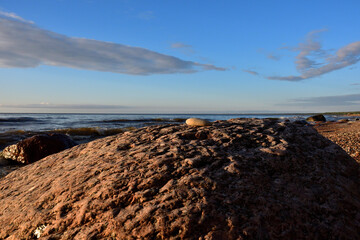 A small light stone on a pink granite boulder in the evening golden sunlight on the shore against the background of the sea with a slight breeze under a blue sky with beautiful light clouds.