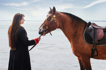 Obraz premium A beautiful woman with long and black hair in a historical hussar costume stands near a river with a horse.
