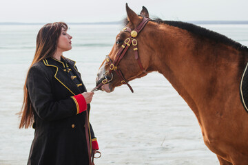 Obraz premium A beautiful woman with long and black hair in a historical hussar costume stands near a river with a horse.