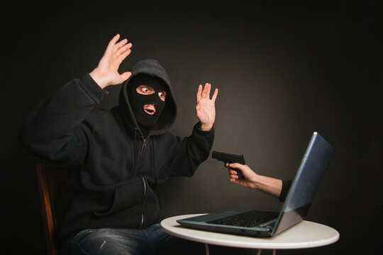 Caught an internet fraudster. photo concept of digital robbery. masked man is afraid of hand with pistol from laptop. studio photo