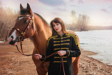Obraz premium A beautiful woman with long and black hair in a historical hussar costume stands near a river with a horse.
