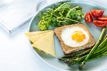 Sunny side up egg in a toast served with cheese, asparagus, rocket and tomatoes on a rustic white background