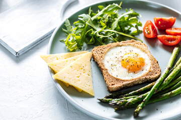 Sunny side up egg in a toast served with cheese, asparagus, rocket and tomatoes on a rustic white background