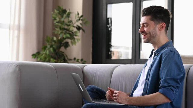 Happy friendly caucasian man sitting on a sofa at home, wearing stylish denim clothes, using a laptop, chatting with friends or family via video conference, waving his hand, smiling