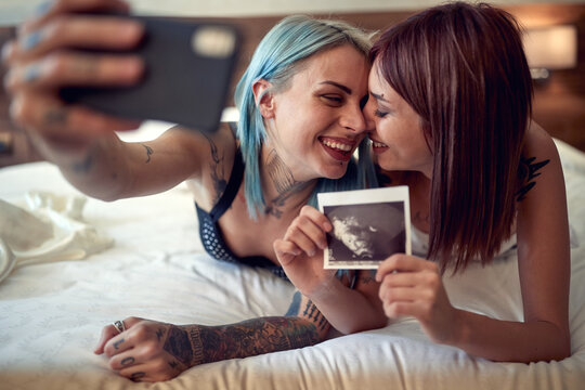 A happy young lesbian couple is lying in the bed full of emotions while taking selfie with ultrasound photo. Home, relationship, love