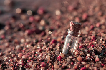 Horizontal close-up photography of a single miniature empty glass chemical bottle closed by cork, stuck into the beach colorful sand