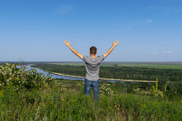 Young man stands on high hill and enjoying natural environment. Happy tourist holds his hands up to the sky and looking at river and green endless fields.