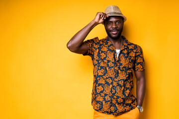 Handsome african american man in colorful shirt holds his hat with his hand on his head. Isolated on a yellow background. Summer concept