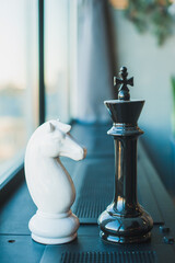 Chess pieces, a white knight and a black queen, large pieces stand on a black windowsill near the window in the room.