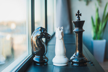 Chess pieces, a black and white knight and a queen, large pieces stand on a black windowsill near the window in the room.