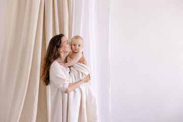 Fototapeta premium Mom hugs baby on a white background. Eco parenting concept. White clothes and a diaper natural fabrics linen cotton muslin. Mother's love. Hugs, smiles, happiness. Mother and child togetherness