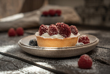 Delicious tartlet with custard, fresh raspberries and sugar powder, served on gray ceramic plate over wooden surface. Summer seasonal sweets. Soft focus.