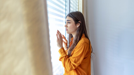 Cropped shot of a beautiful woman looking out the window at home. Sad young woman suffering from agoraphobia looking out of window. Depressed woman standing by window at home. Depressed woman at home. - obrazy, fototapety, plakaty