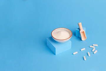 Collagen protein powder in a bowl and capsules on a blue background. A food supplement for the skin and joints. Creative photo.